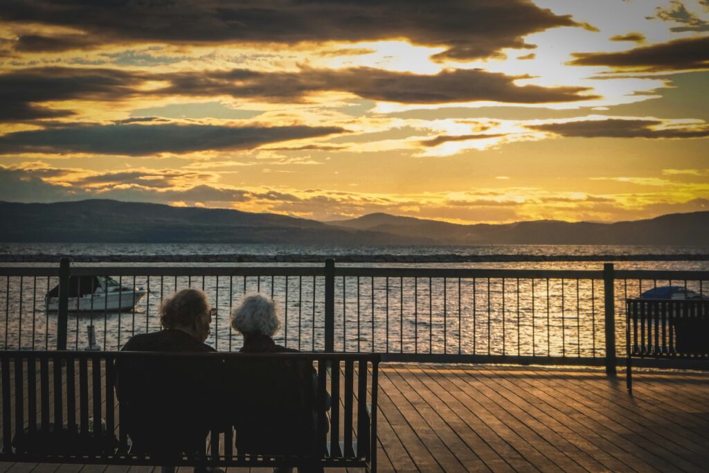 Retiring Abroad-an older couple sitting on a bench by the shore watching the sunset.