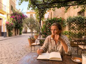 living abroad-an older woman sitting in an outdoor cafe in Spain, sipping coffee while writing in a notebook